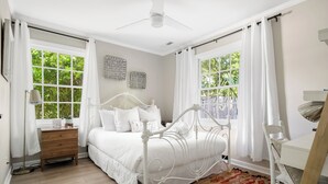 Ein Schlafzimmer mit einem weißen Bett, einem Holfnachtkästchen, einem Schreibtisch und einem gemusterten Teppich.