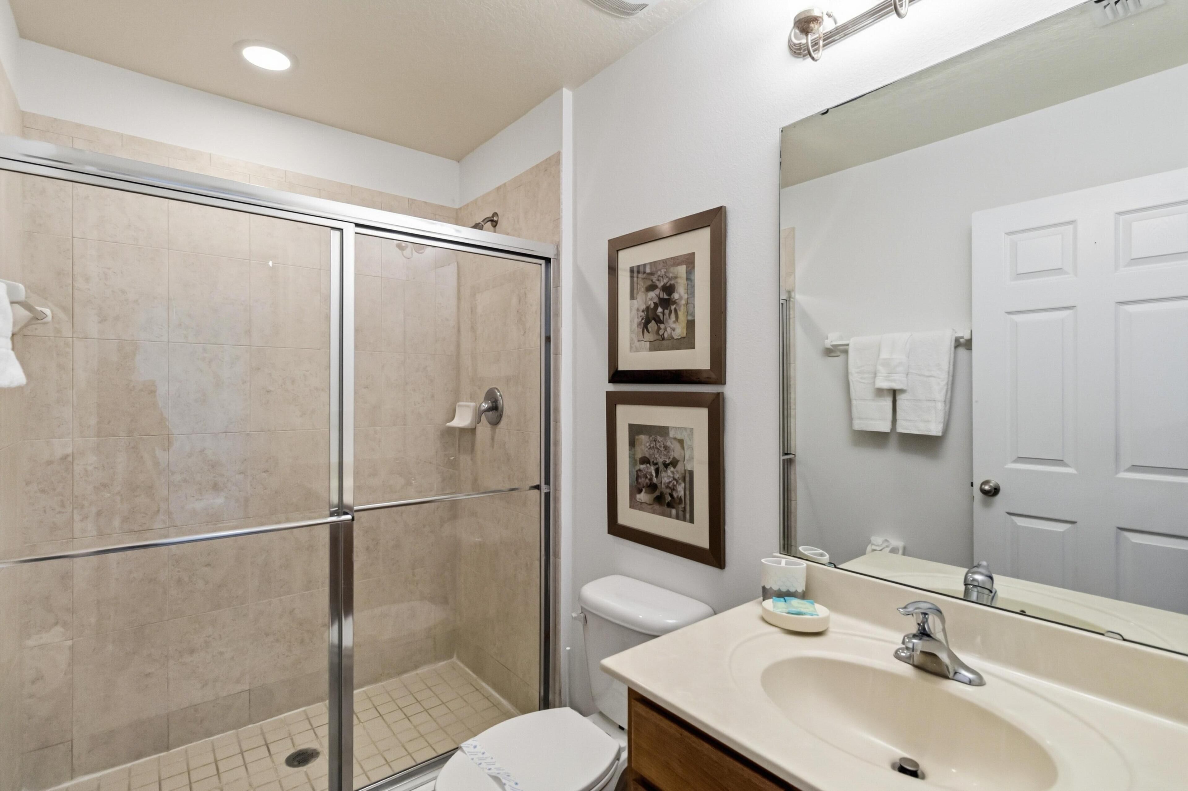 Un baño con cabina de ducha de vidrio, inodoro, lavabo con espejo y dos cuadros enmarcados en la pared.