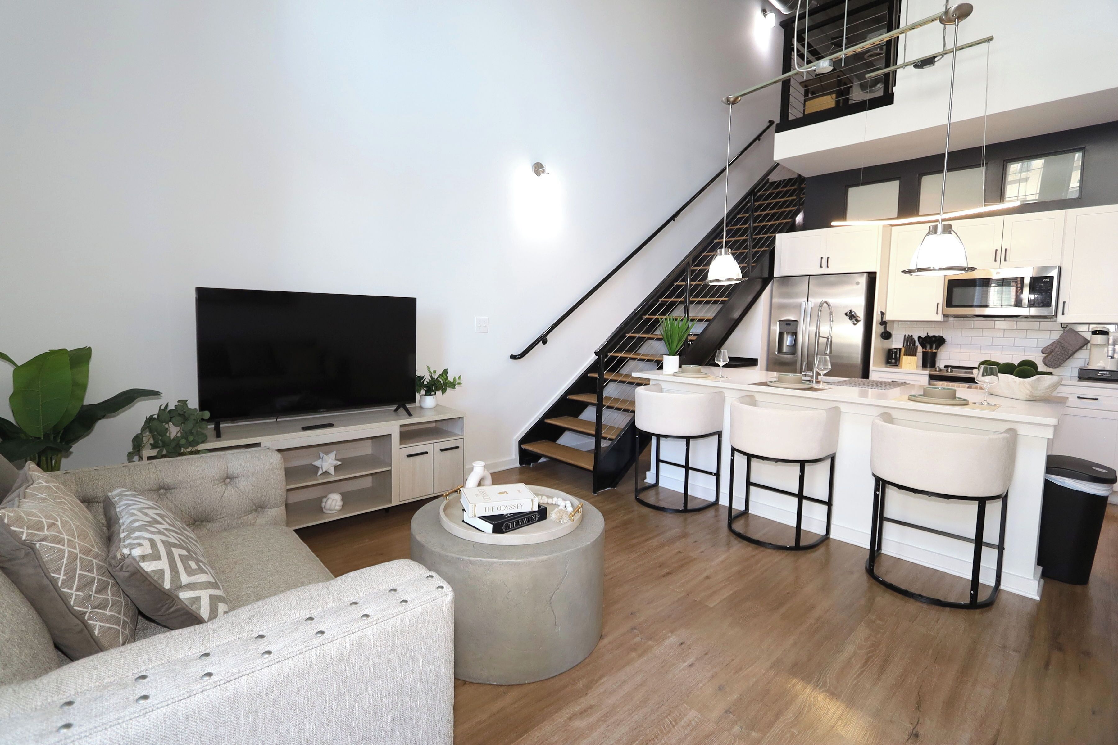 A modern living room with a flat-screen TV, a sofa, a coffee table, and a kitchen area with a microwave, refrigerator, and bar stools.