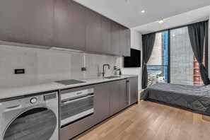 Eine moderne Küche mit integriertem Wascher, Backofen und Spüle, ein großes Fenster mit Vorhängen sowie ein Bett im angrenzenden Raum.