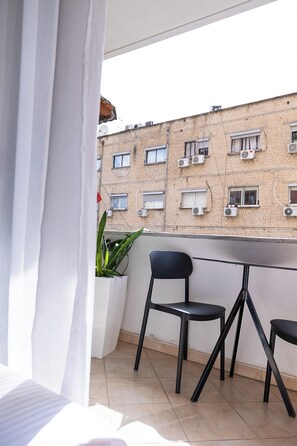 Ein Balkon mit Blick auf ein Backsteingebäude, zwei schwarze Stühle, ein Blumentopf und ein weißer Vorhang.
