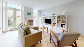 A living room with a sofa, armchair, TV, bookshelf, and a door to a balcony. - Bright 2BR Home w/ Private Garden Near Wimbledon (London)