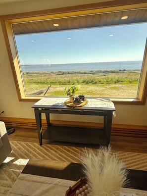 A room with a large window overlooking a grassy field and the sea, a small table with a plant and a decorative item, and a chair with a patterned cushion.