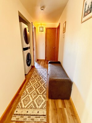 Un pasillo con lavadora, alfombra estampada, banco y una puerta.