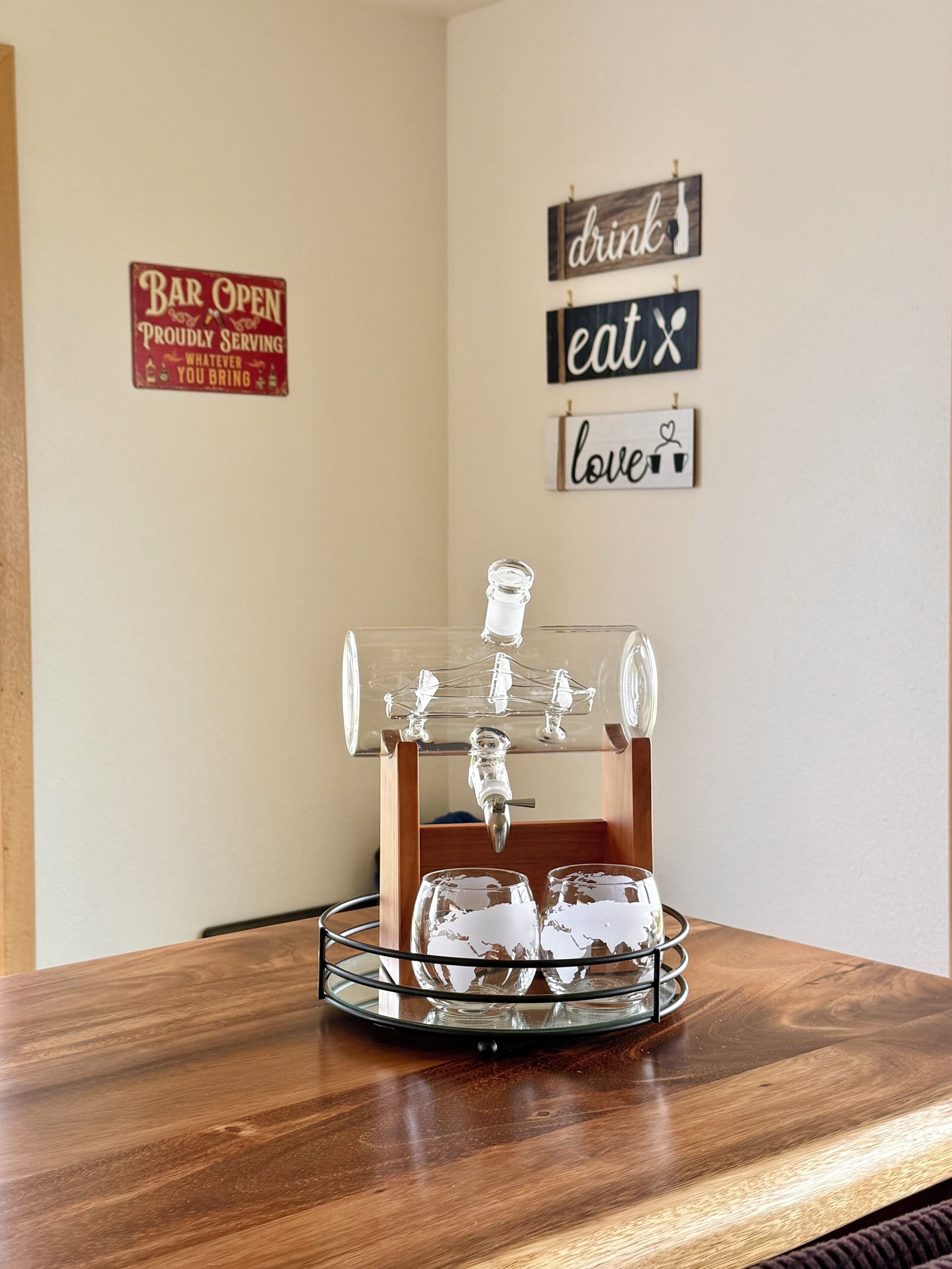 Una barra con grifo y vasos sobre una mesa de madera, rodeada de carteles en la pared con palabras como 'beber', 'comer' y 'amor'.