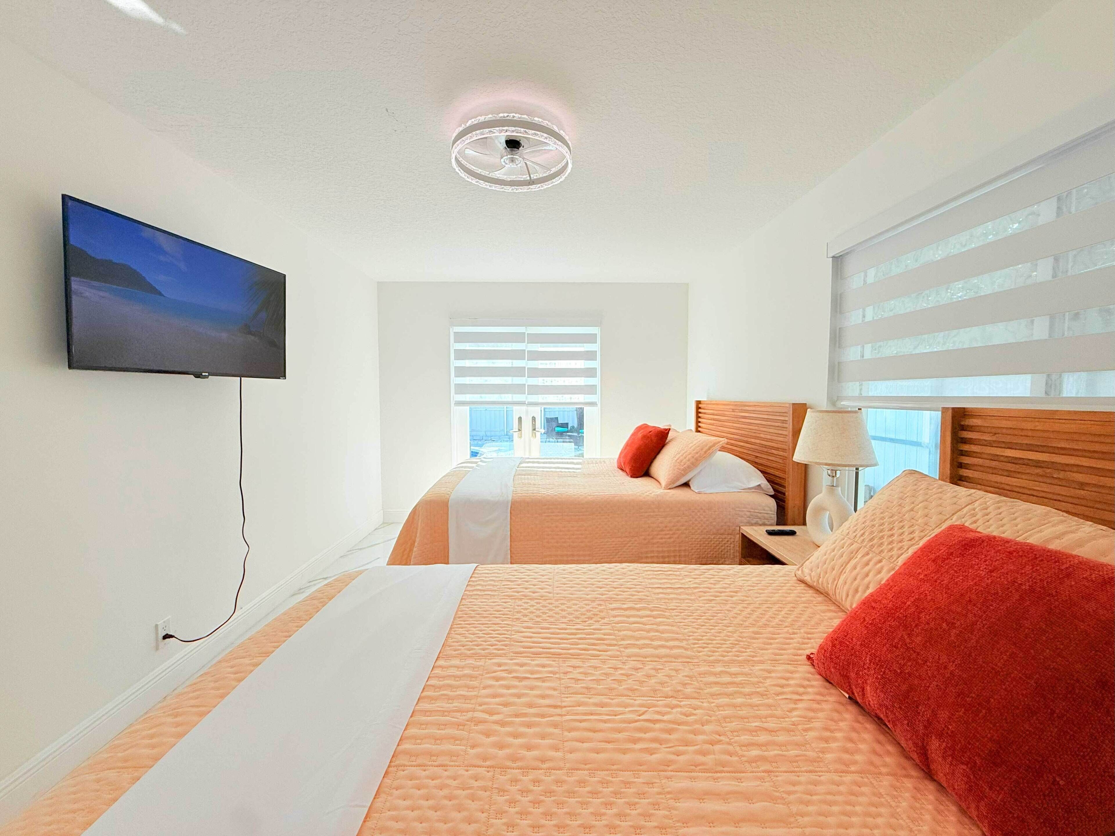 A hotel room with two beds, a television, a ceiling fan, and a window with blinds.