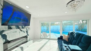 Ein geräumiges Wohnzimmer mit einem großen Flachbildfernseher an einer Marmorwand, einer modernen Kristallleuchtre und Blick auf einen Swimmingpool durch große Schiebeglastüren.