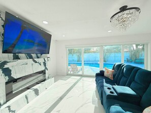 A spacious living room with a large flat-screen TV mounted on a marble wall, a modern chandelier, and a view of a swimming pool through large sliding glass doors. - Private Pool, Arcade Games & Family Fun Near Beach (Fort Lauderdale)