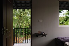 Ein Zimmer mit Balkon, einem kleinen Regal mit einem Buch, einem Fenster mit Blick auf Grünflächen und einem Bett mit violetter Bettdecke.
