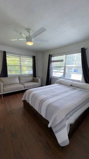 Ein Schlafzimmer mit einem Bett, einer Couch, einem Deckenventilator und großen Fenstern mit Vorhängen.