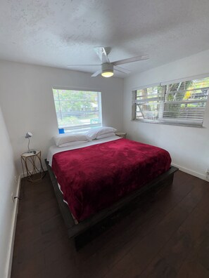 Une chambre à coucher comprenant un lit, un ventilateur de plafond, une fenêtre avec des stores et une petite table de chevet.