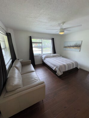 Une pièce comprenant un lit, un canapé, un ventilateur de plafond, une fenêtre avec des rideaux et un tableau accroché au mur.