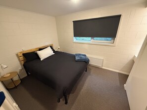 Un dormitorio con cama, ventana con persianas, radiador y mesita de noche pequeña.