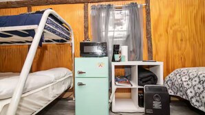 Uma cama de solteiro, uma pequena geladeira, um café, uma prateleira com livros e uma cama com roupa de cama estampada.