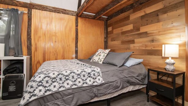 Quarto com teto de madeira, cama com manta estampada, almofadas, mesa de cabeceira com abajur e prateleira pequena com um laptop.