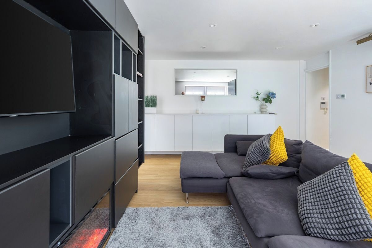 A modern living room with a grey sectional sofa, a flat-screen TV, and a kitchen in the background.