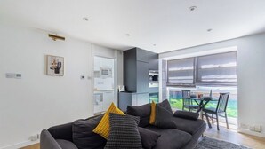 A modern living room with a dark-colored sofa, a dining area with a table and chairs, and a view through large windows.