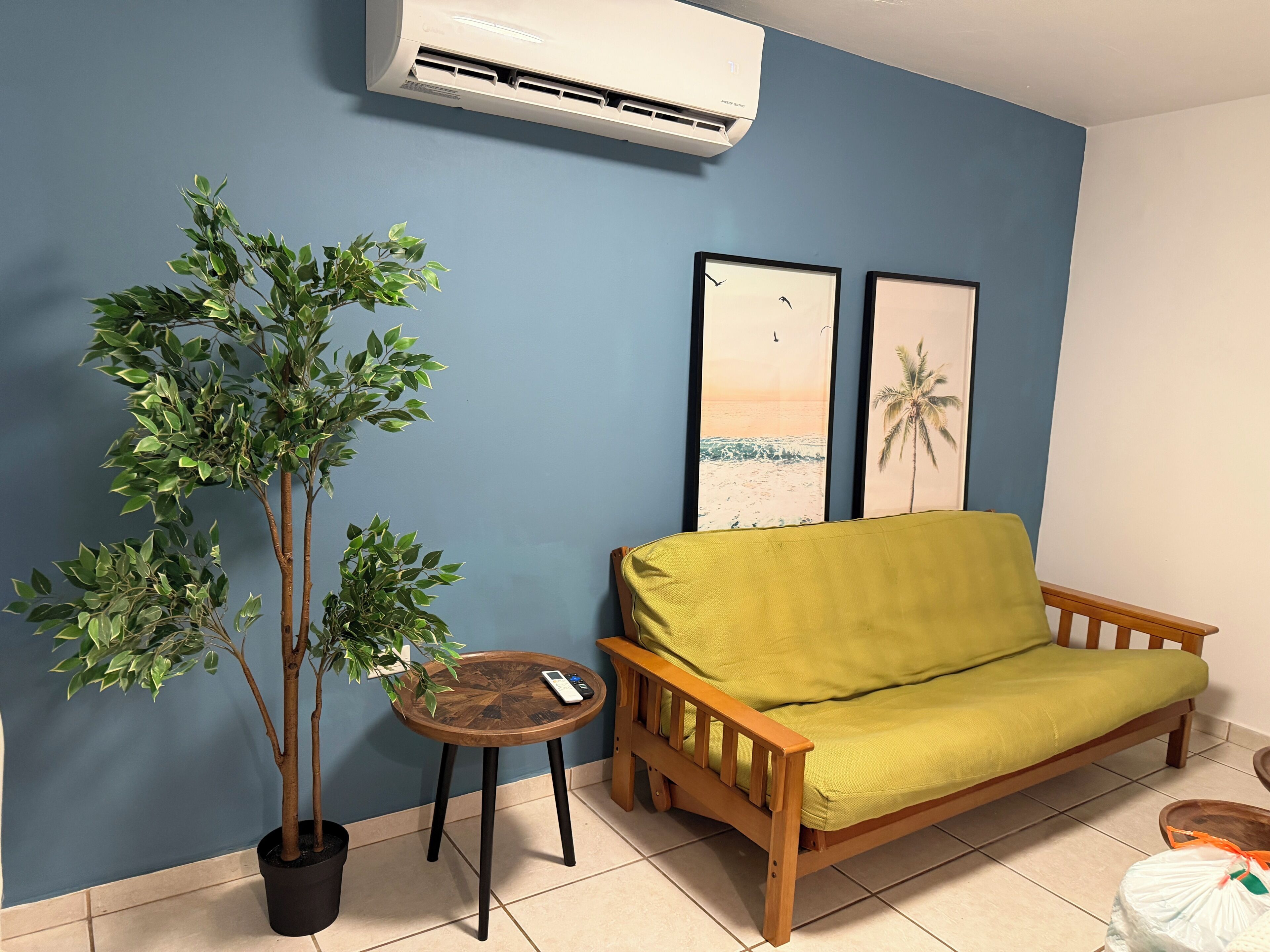 Ein Zimmer mit blauer Wand, einem Holzsofa, einem kleinen runden Tisch mit Fernbedienung, einer Topfpflanze und zwei gerahmten Bildern.