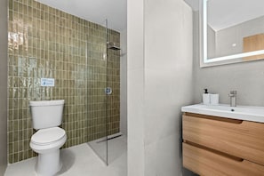 A bathroom with a toilet, shower, and vanity with a mirror. - Villa Playa - Beach & Pool Apartments (San Juan)