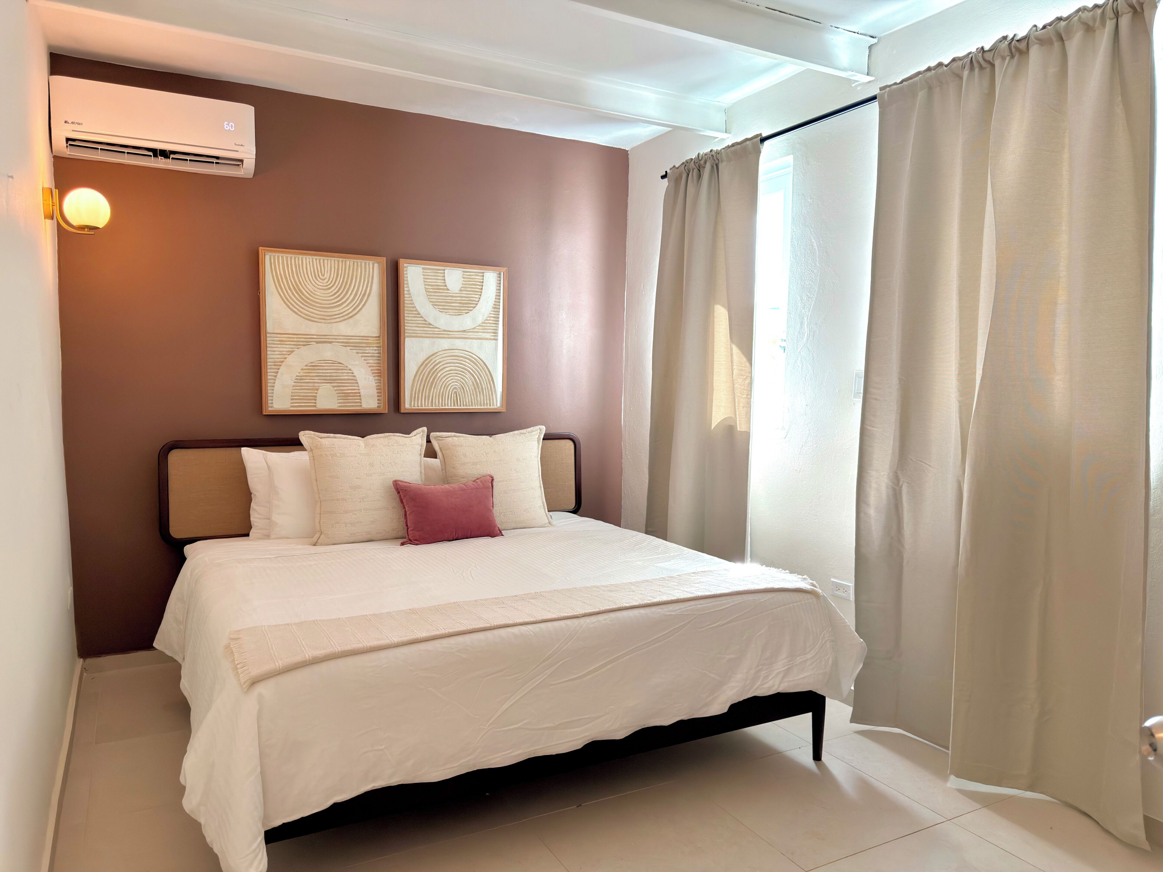 A bedroom with a bed, pillows, a wall-mounted air conditioner, and two abstract paintings.