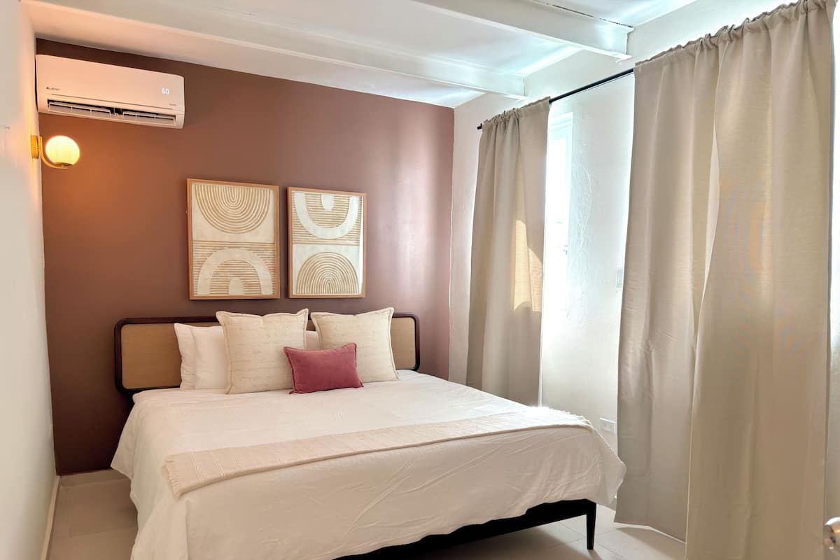 Un dormitorio con cama, almohadas, aire acondicionado de pared y dos cuadros abstractos.