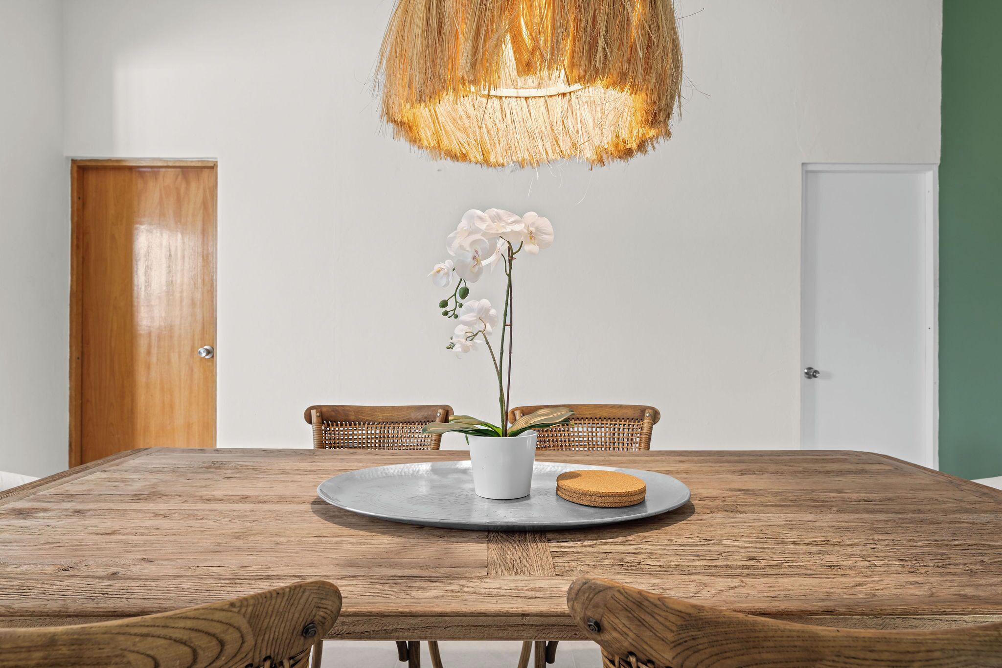 Una mesa de comedor de madera con un jarrón blanco lleno de orquídeas blancas, dos sillas de mimbre y una lámpara colgante de fibras naturales.