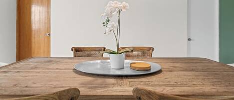 Una mesa de comedor de madera con un jarrón blanco lleno de orquídeas blancas, dos sillas de mimbre y una lámpara colgante de fibras naturales.