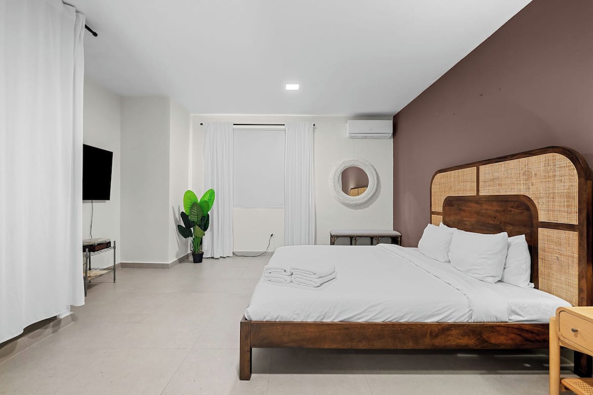 Un dormitorio con cama de madera, ropa de cama blanca, una mesita de noche, una planta en maceta, un espejo redondo y un televisor en la pared.