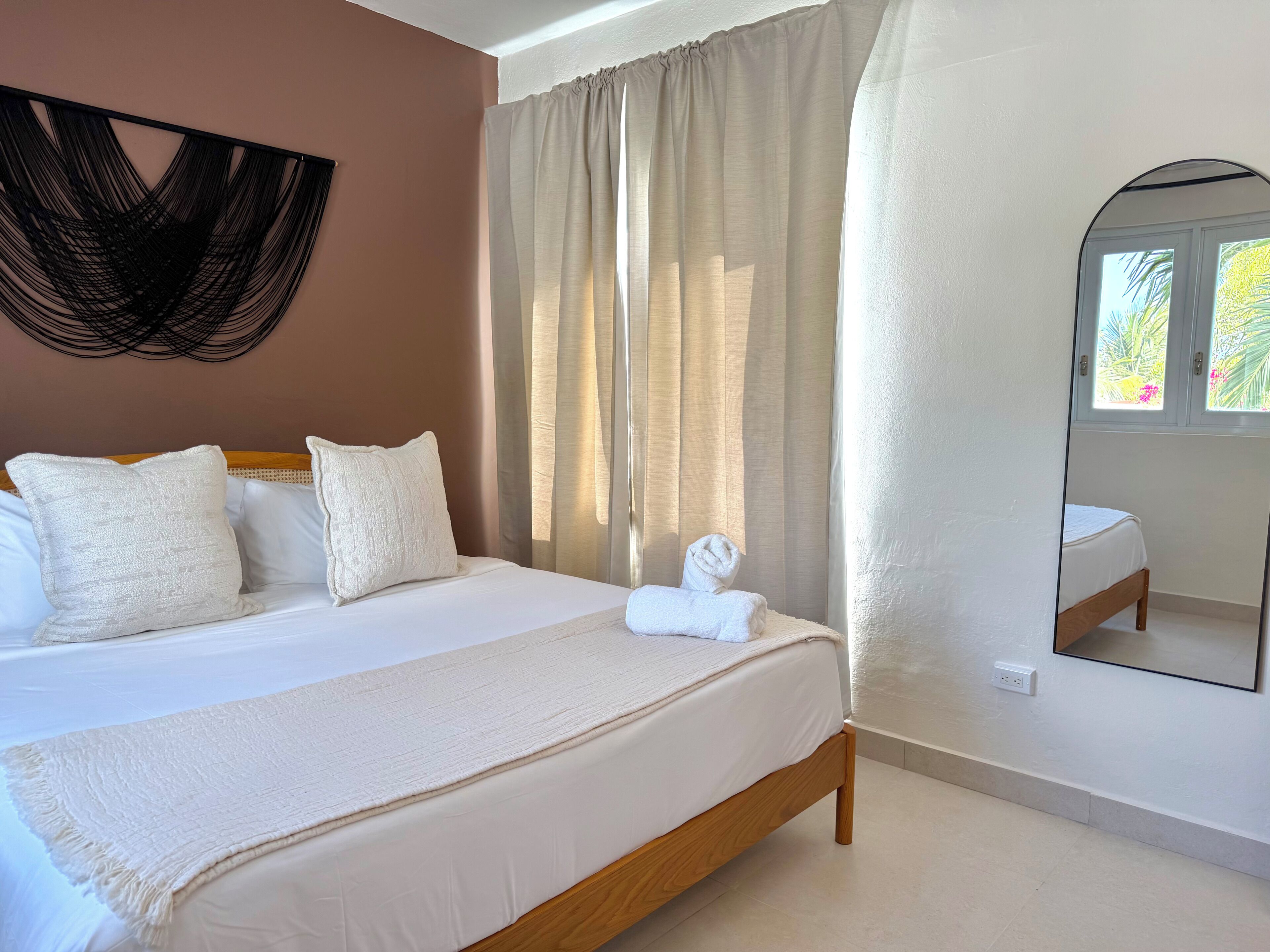 A bedroom with a bed, pillows, a bedside table, a mirror, and a window with curtains.