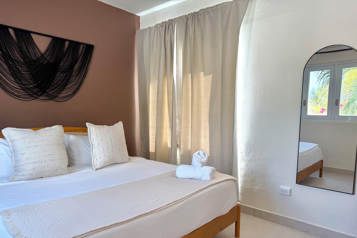Un dormitorio con cama, almohadas, una mesita de noche, un espejo y una ventana con cortinas.