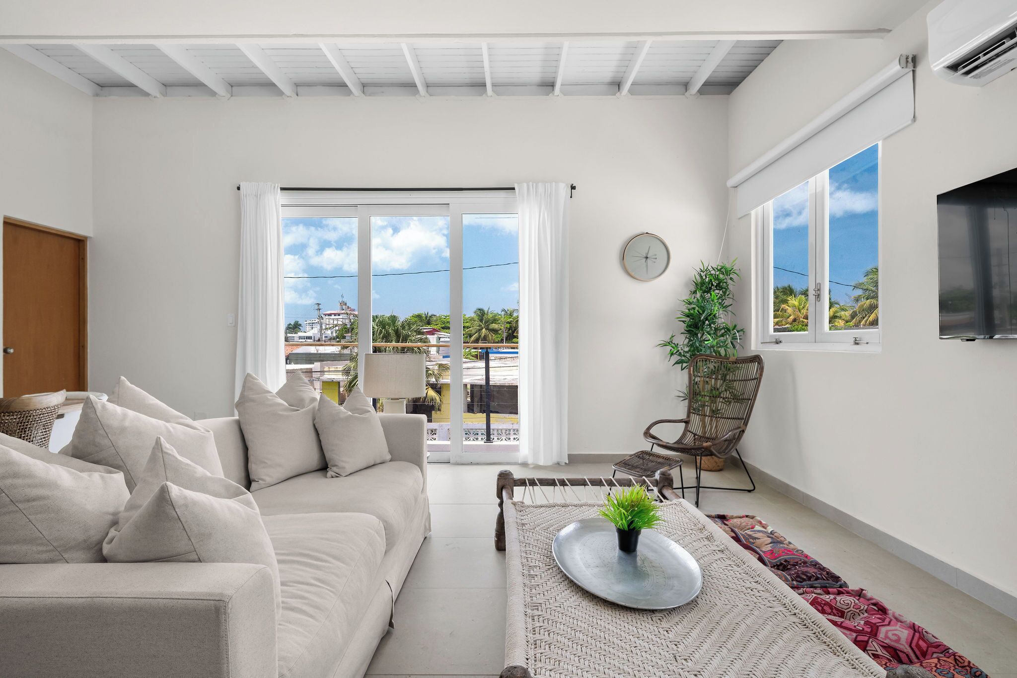 A modern living room with a sofa, a coffee table with a plant, a chair, a clock, and a view through a sliding door to the outside.