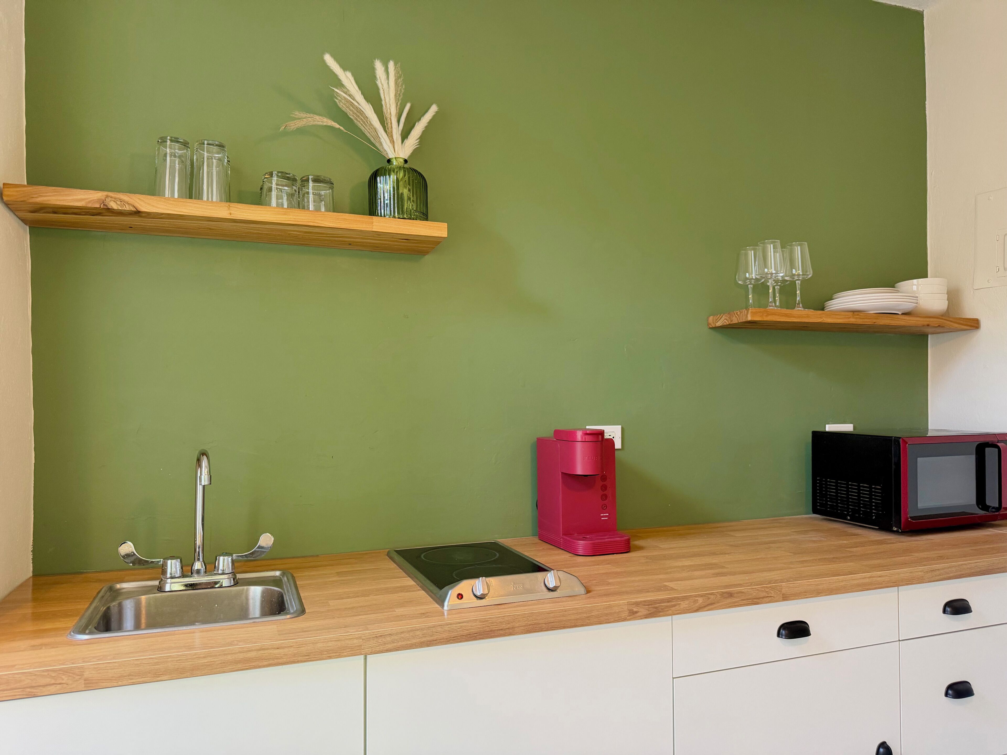A kitchen with a sink, a toaster, a red coffee maker, a microwave, and shelves with glass jars and a vase.