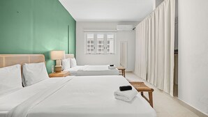 Ein Hotelzimmer mit zwei Betten, einem Fenster mit Rollläden, einem Nachttisch mit Lampe und einem durch einen Vorhang einsehbarer Badezimmer.