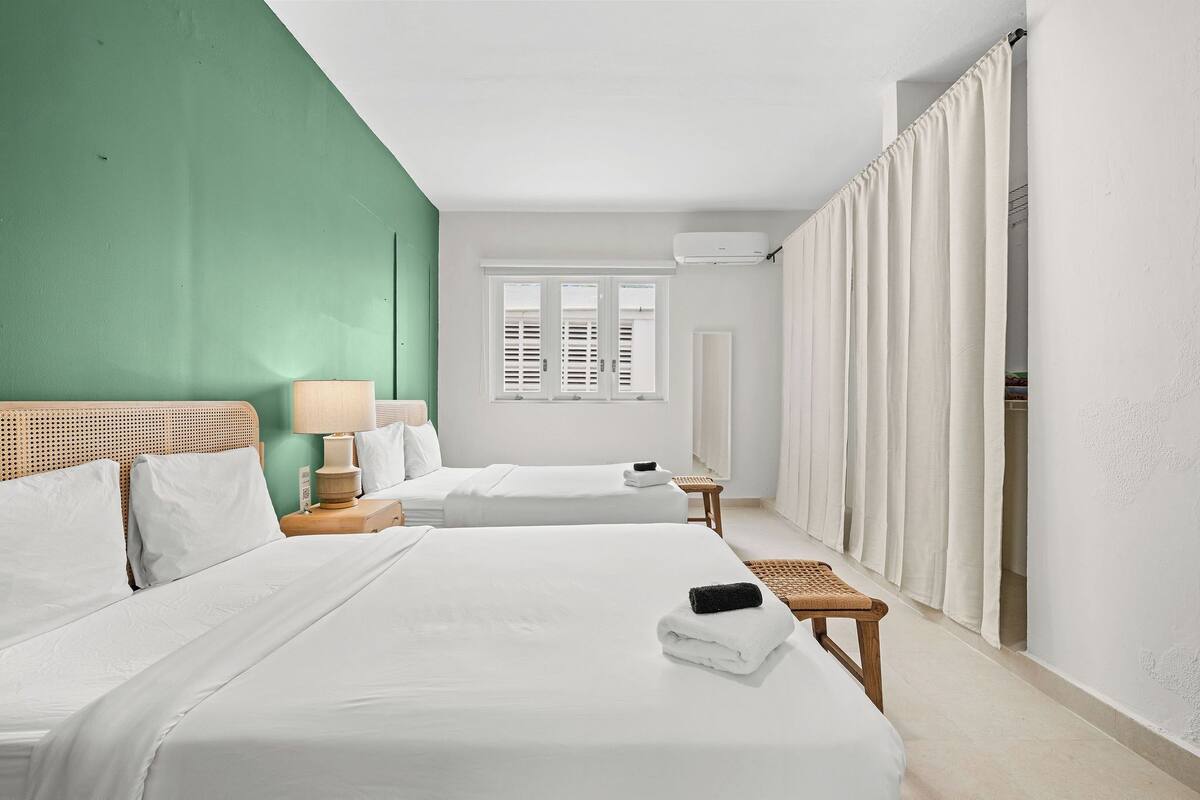 Habitación de hotel con dos camas, una ventana con contraventanas, una mesita de noche con lámpara y un baño visible tras una cortina.