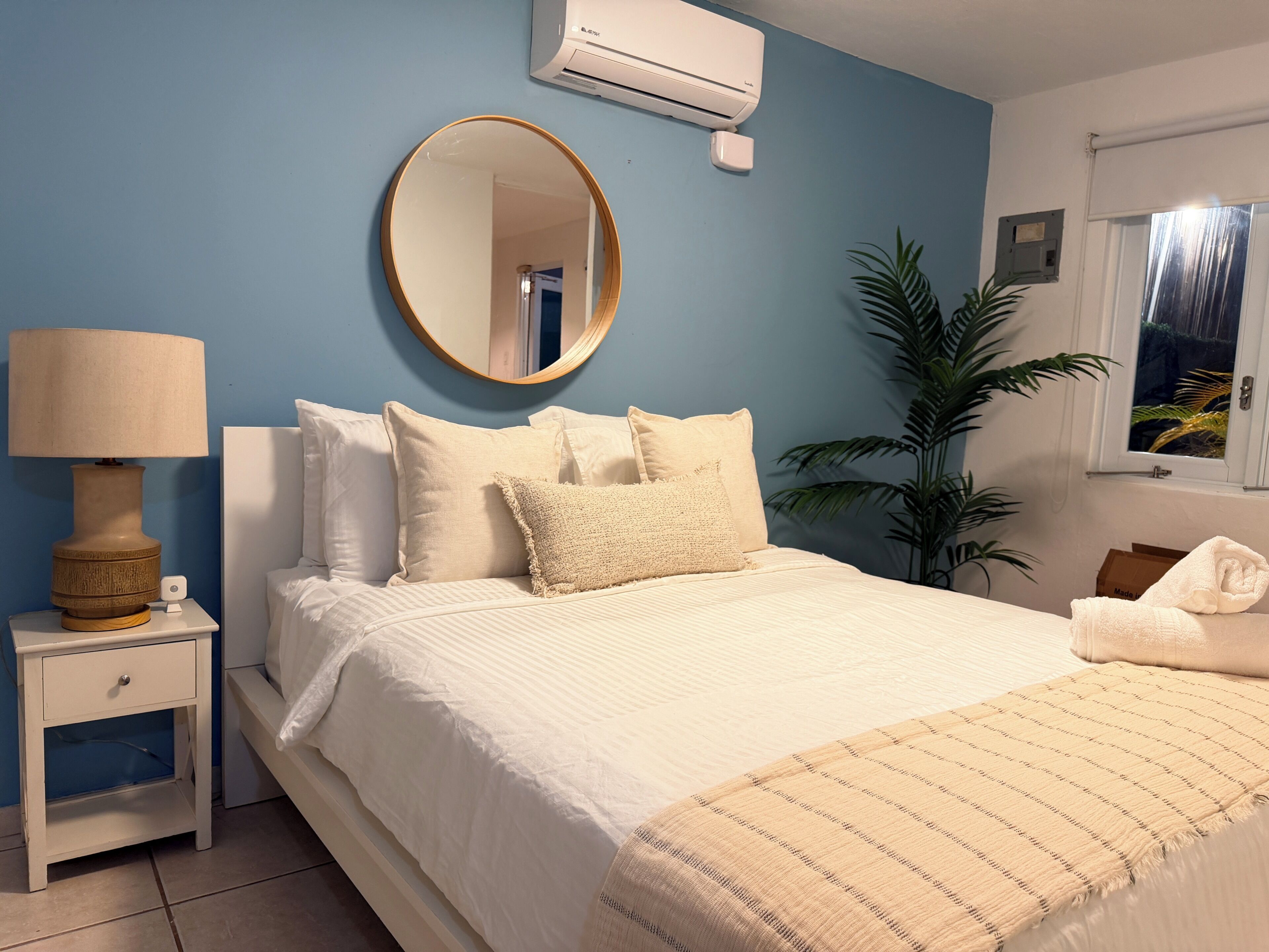 A bedroom with a bed, bedside table, lamp, mirror, air conditioning unit, potted plant, and a window.