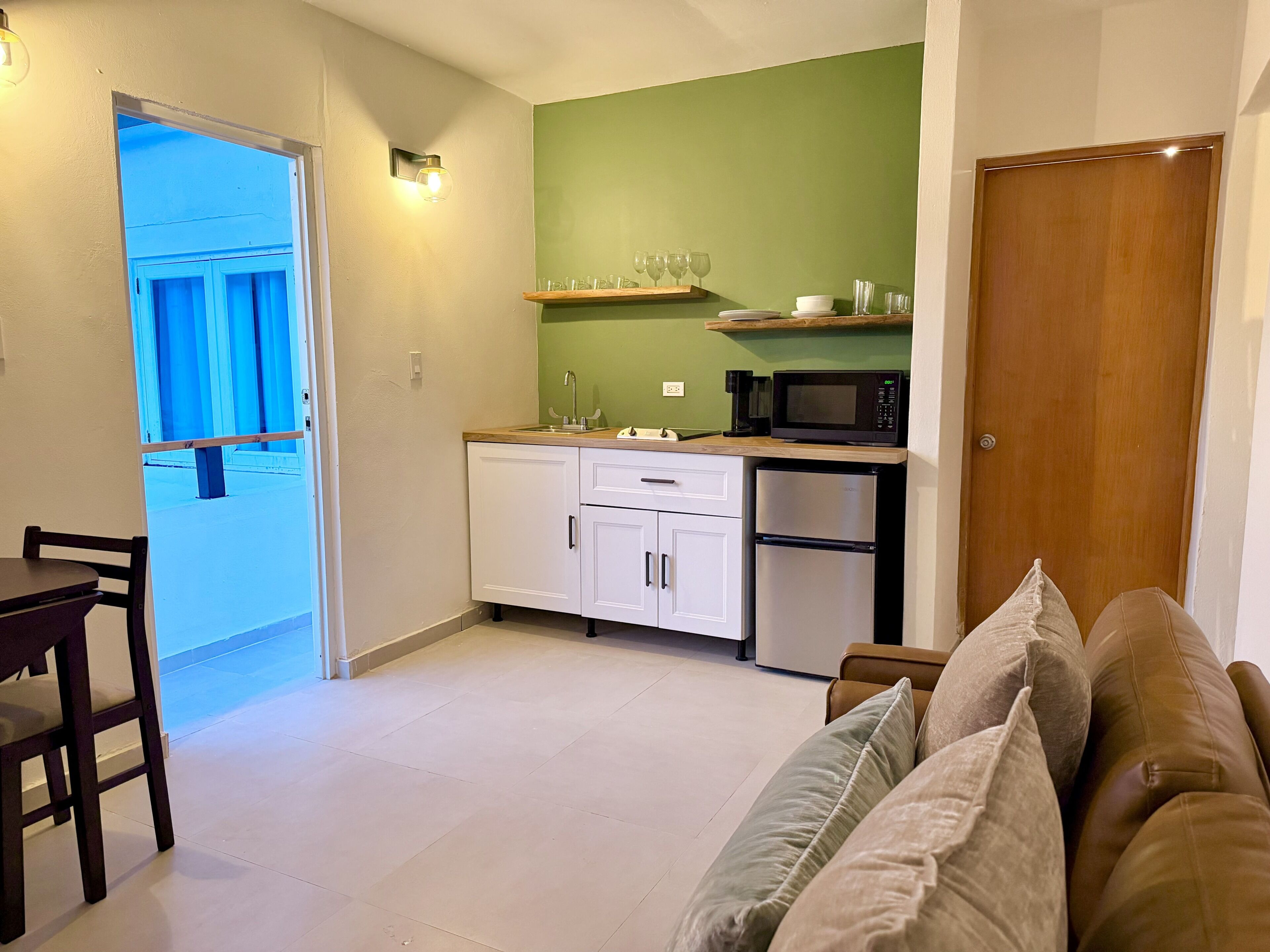 A compact kitchen with a microwave, a coffee maker, and a small refrigerator. There is a dining table with chairs and a sofa with cushions.