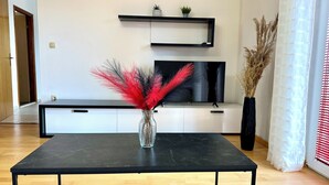 A modern living room with a black coffee table, a flat-screen TV, white cabinetry, and decorative plants. - Magic Sunset – apartman za 4 osobe s velikim balkonom i orbitrekom (Pirovac)