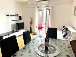 Un saló modern amb una taula de dinar preparada per a dues persones, un televisió pantalla plana, un sofà i una finestra amb persianes.