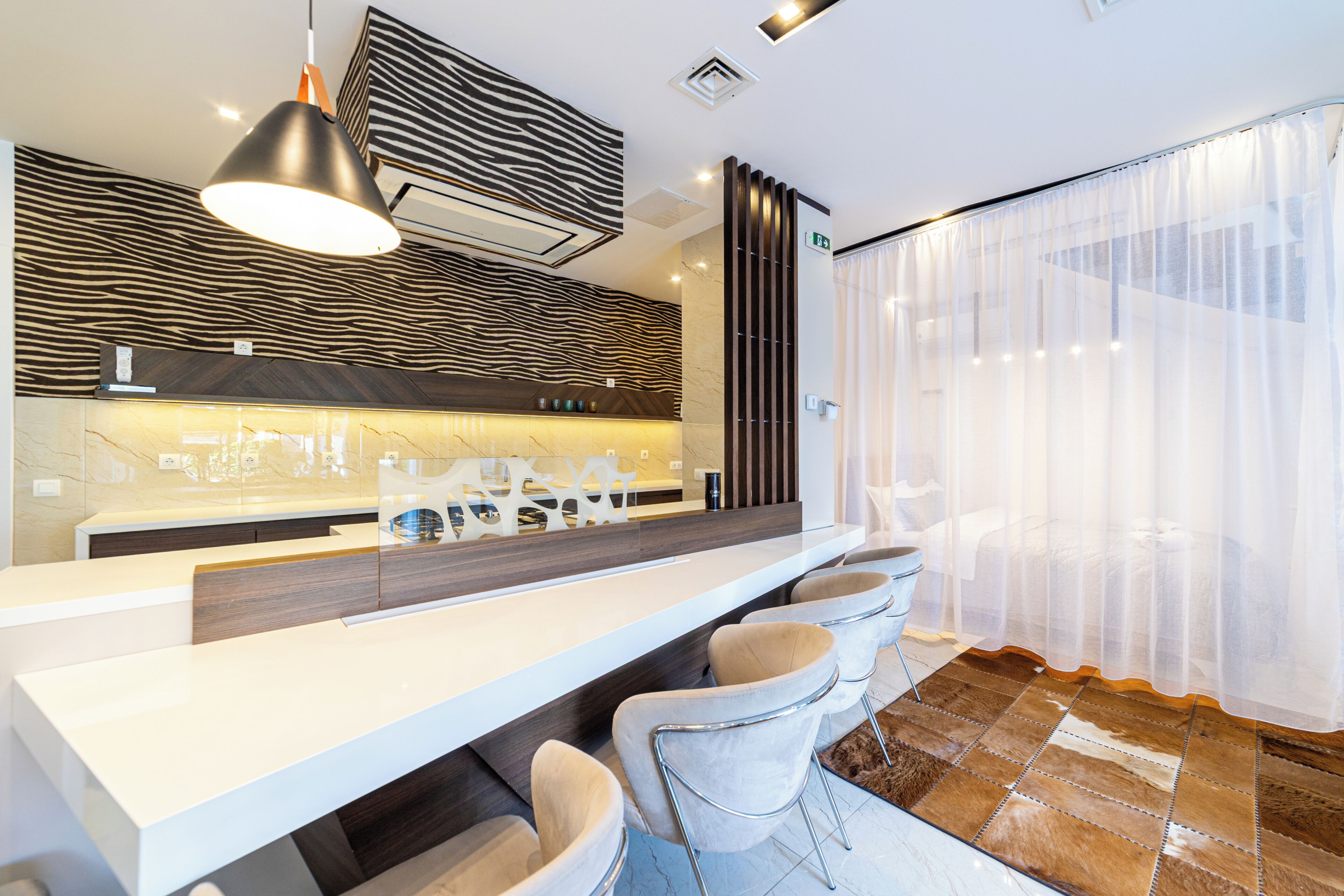 Un espace bar moderne avec un comptoir blanc, des tabourets de bar en cuir, un mur à motifs zébrés et un grand luminaire suspendu.
