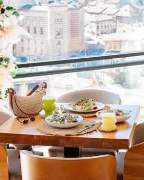 Una mesa con platos de comida, un bolso tejido y una bebida, con vista a la ciudad.
