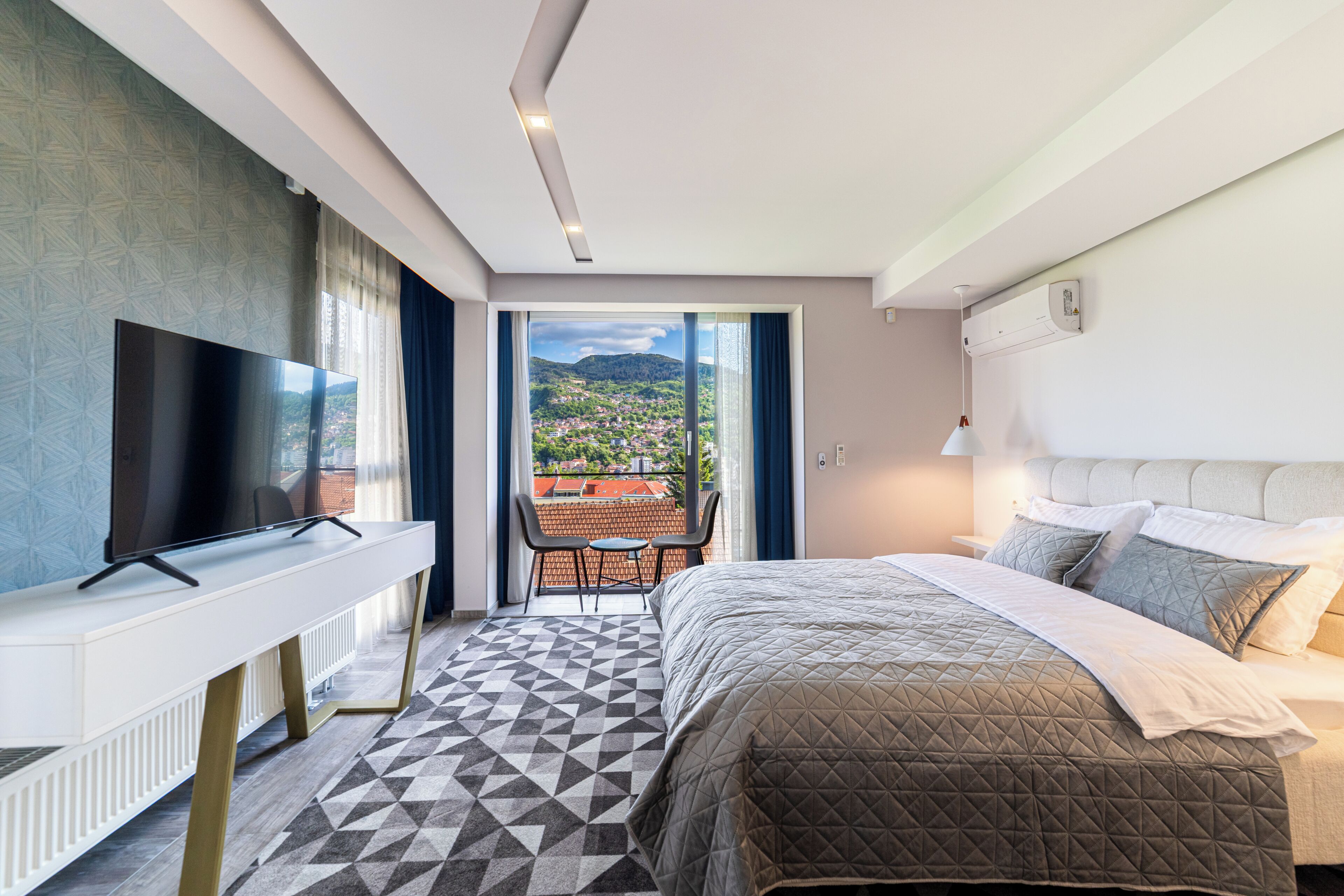 A hotel room with a bed, a TV, a desk, and a balcony with a view of the city.