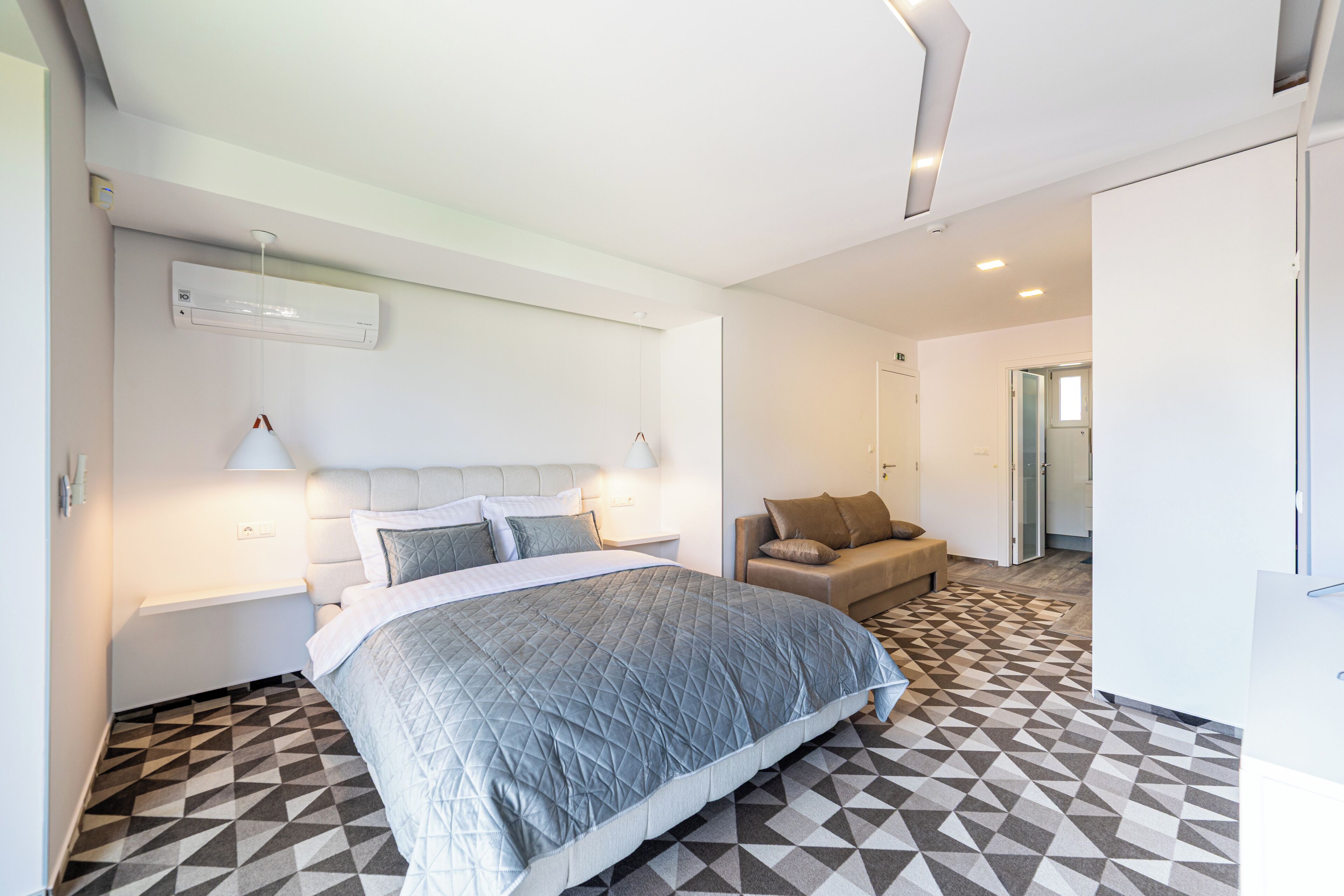 A hotel room with a bed, a sofa, a wall-mounted air conditioner, and a patterned floor.