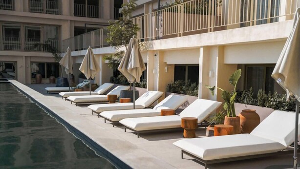 Zona junto a la piscina con tumbonas y sombrillas, una piscina cristalina y un edificio con balcones al fondo.
