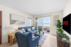 Ein Wohnzimmer mit Sofa, Couchtisch und Fernseher, mit Blick auf den Strand durch eine Schiebetür.
