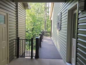 Un couloir extérieur abrité, doté d’une terrasse en bois, d’une rambarde en métal et offrant une vue sur un espace verdoyant et arboré.