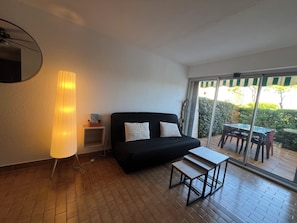 Une pièce avec un canapé noir, une petite table, une lampe et une porte-fenêtre coulissante donnant sur un espace extérieur équipé d’une table et de chaises.