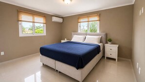 A bedroom with a bed, bedside tables, a window with blinds, an air conditioning unit, and a view of greenery outside. - Tropical 3-Bedroom Villa with Pool Next to the Jan Thiel Salt Flats (Willemstad)
