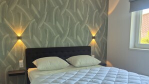 Una cama con edredón blanco acolchado, cabecera oscura y dos lámparas fijadas a la pared.
