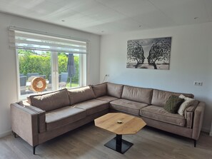 Un salon moderne avec un canapé d’angle, une table basse en bois et une grande fenêtre équipée de stores.