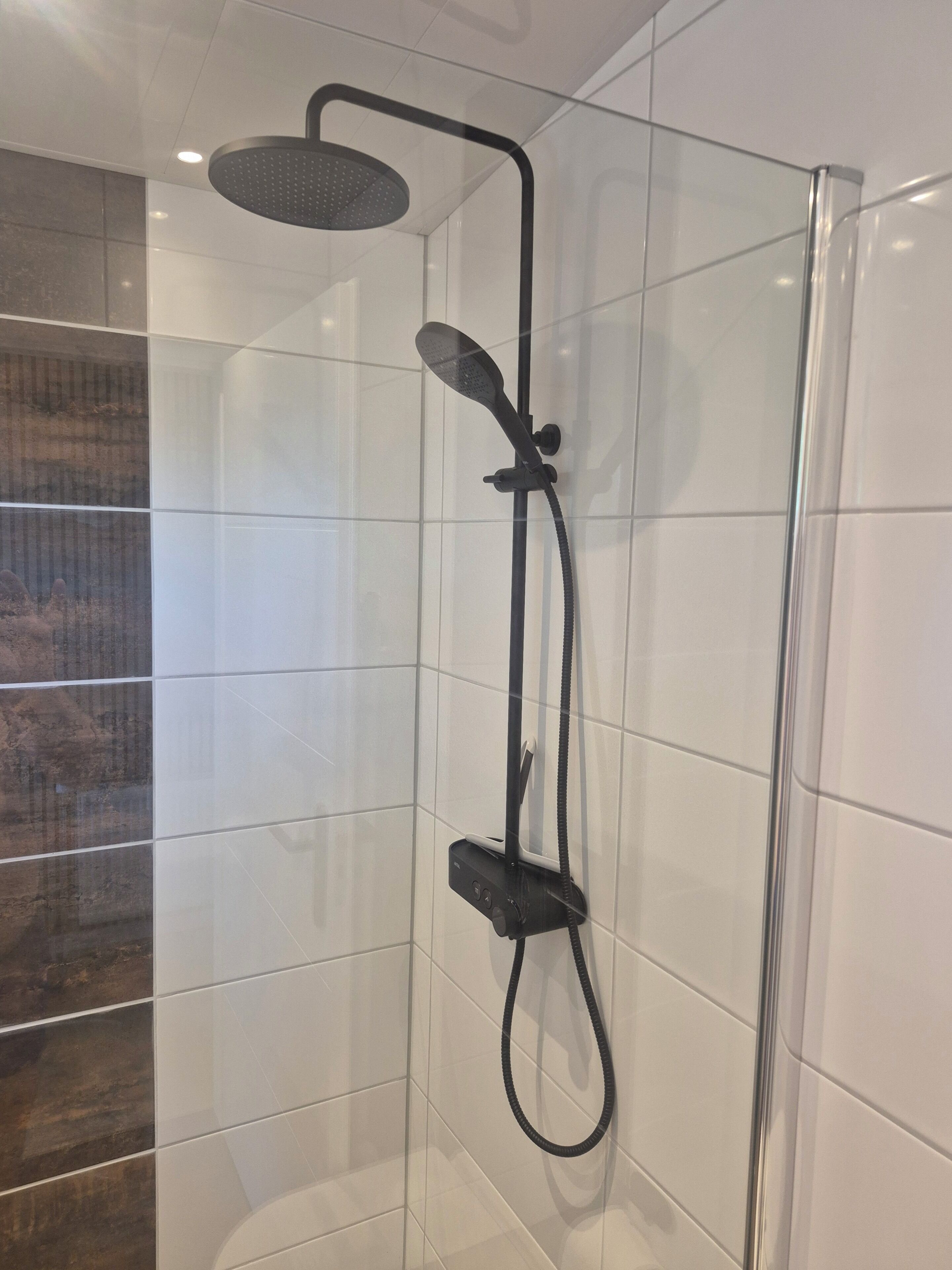 Une douche équipée d’un pommeau de douche à main et d’une douchette à pluie fixée au mur, lequel est recouvert de carreaux.